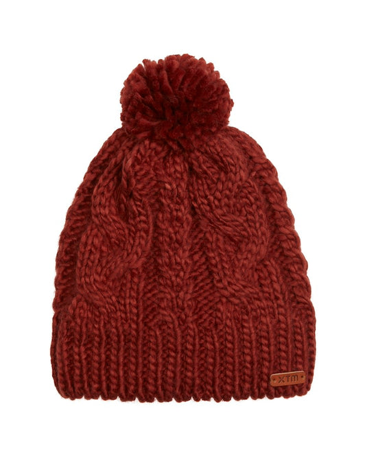 Rust red chunky cable knit XTM Chrystal Beanie with a matching pom-pom and small brown brand tag on the hem.
